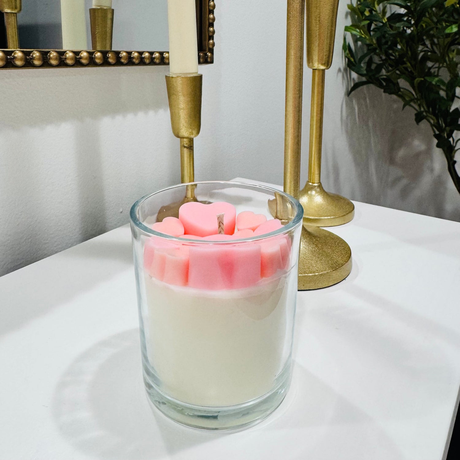 Glass candle holder with pink candles on a white surface, gold candlesticks in the background.